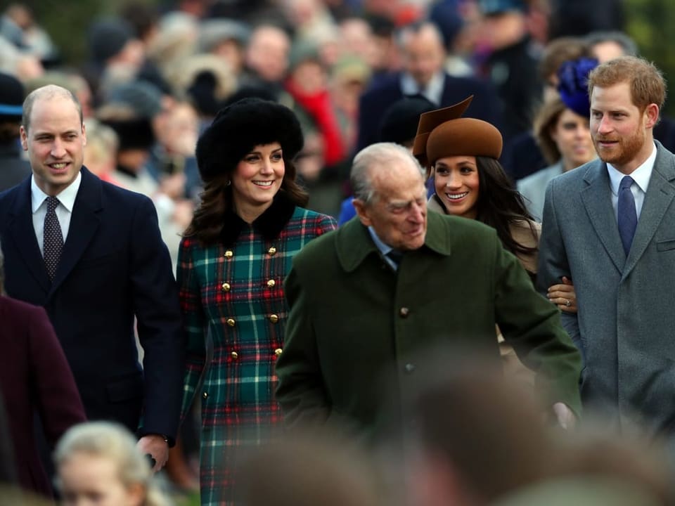 Prinz William, Herzogin Kathrine, Prinz Philip, Meghan und Prinz Harry gehen lächelnd eine Strasse entlang.