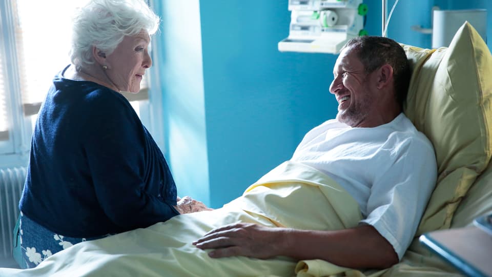 Alte Frau sitzt im Spital am Bettrand, im Bett ein Mann.