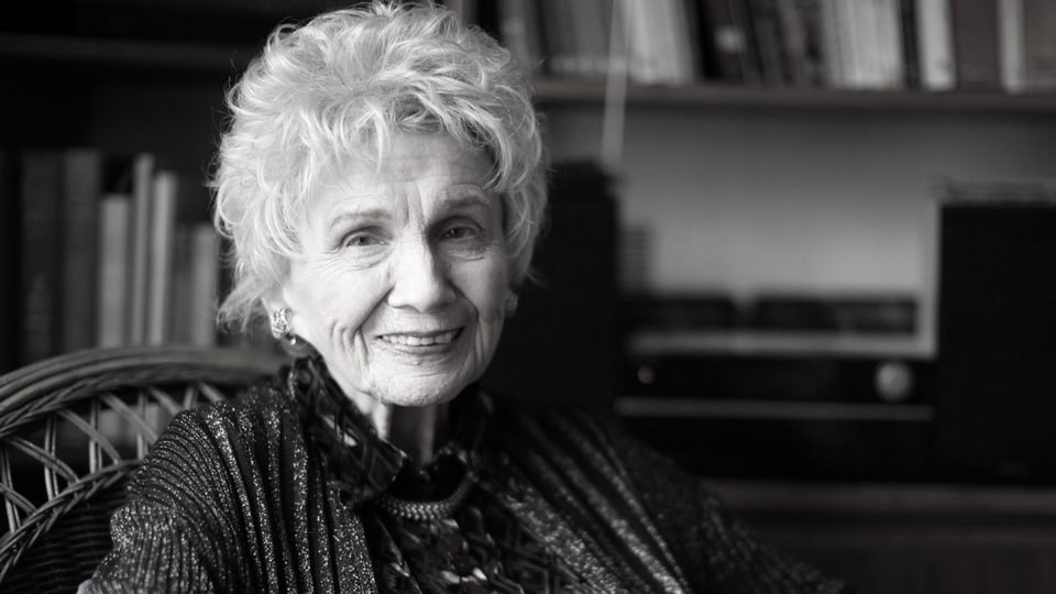 Schwarzweissfoto einer älteren Frau, die lächelt und vor einem Bücherregal sitzt.