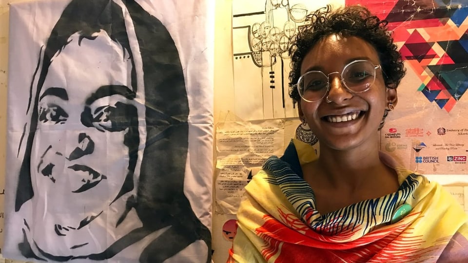 Eine junge Frau mit Brille lächelt neben einem auf Tuch gespraytem Porträt einer Frau mit Kopftuch.