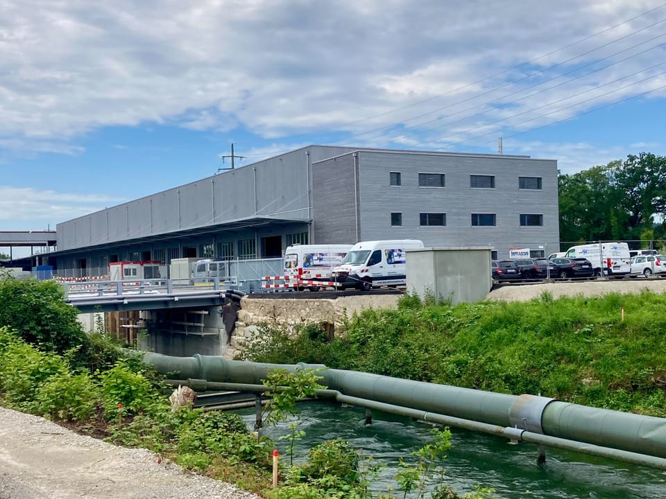 Grünes Wasser und Rohrleitung vor einem grauen Industriegebäude mit geparkten Lieferwagen.
