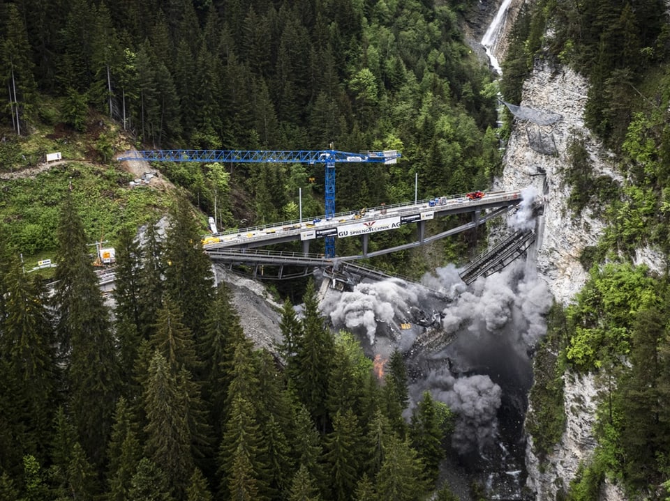 Brücke in einem bewaldeten Tal wird gesprengt, blauer Kran und Staubwolken.