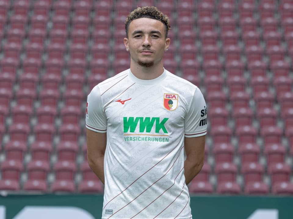 Fussballspieler im weissen Trikot des FC Augsburg steht vor leeren Tribünen.