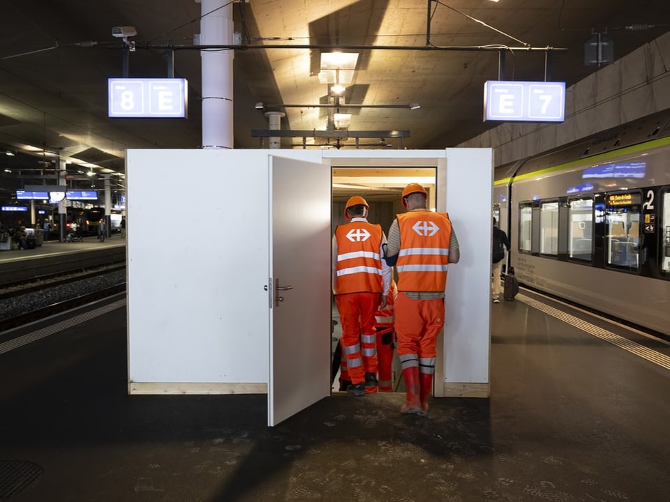 Zwei Arbeiter in orangener Uniform auf Bahnsteig vor weissem Schrank.