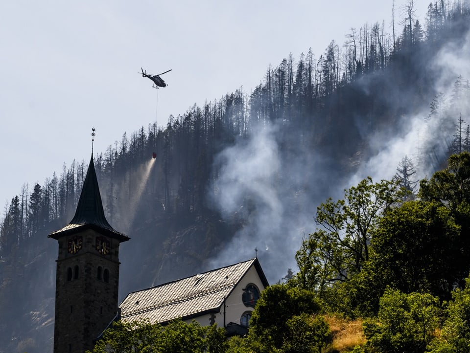 Ein Helikopter wirft Wasser über einem bewaldeten Hang ab. Im Vordergrund steht eine Kirche.