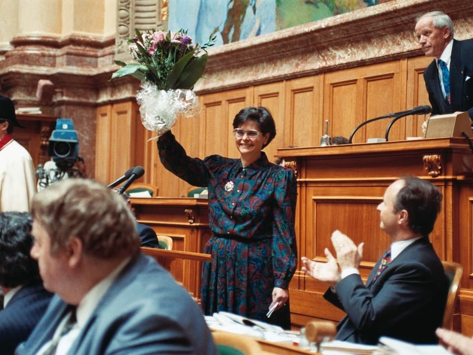 Ruth Dreifuss hält einen Blumenstrauss in die Höhe, ein Mann daneben applaudiert.