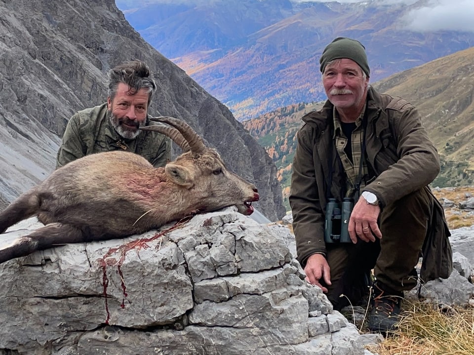 Zwei Männer posieren mit erlegtem Steinbock in Berglandschaft.