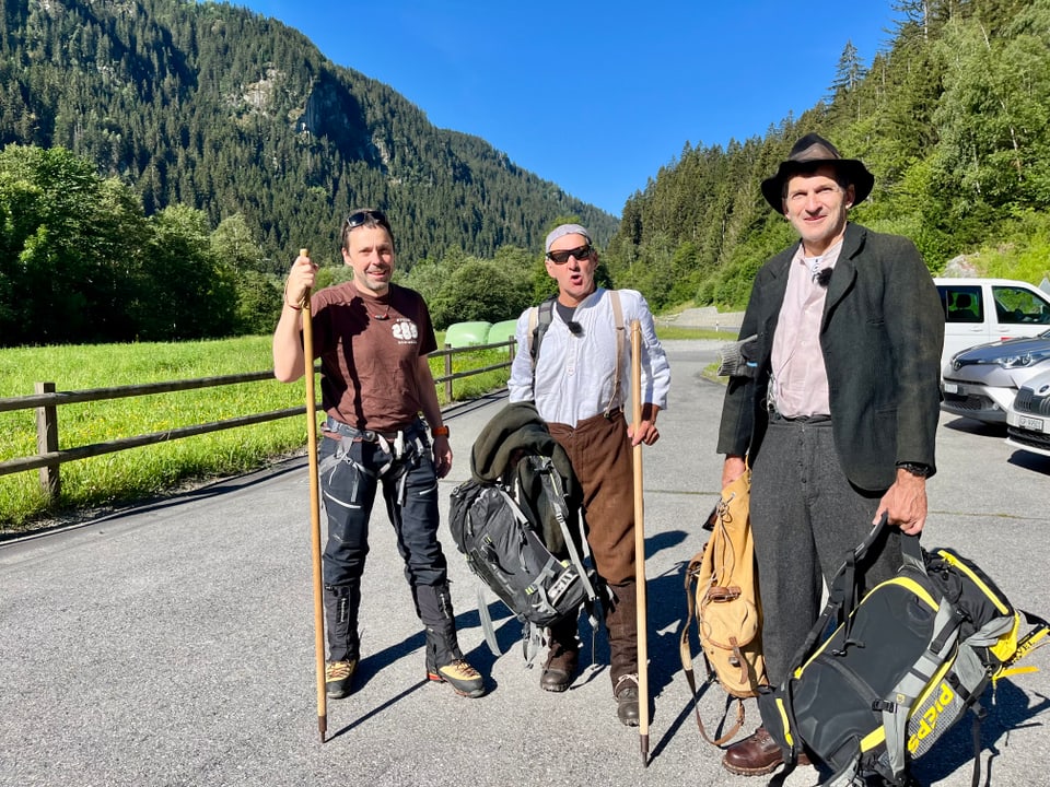 Drei Männer mit Wanderstöcken stehen auf einem Parkplatz vor einer bewaldeten Berglandschaft.