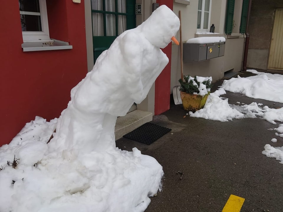 Verschneiter Schneemann, der sich nach vorne lehnt, vor rotem Haus.