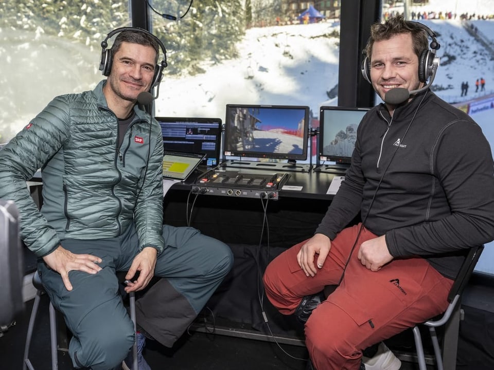 Zwei Männer mit Headsets im Kommentatorraum mit Bildschirmen, verschneiter Hintergrund.