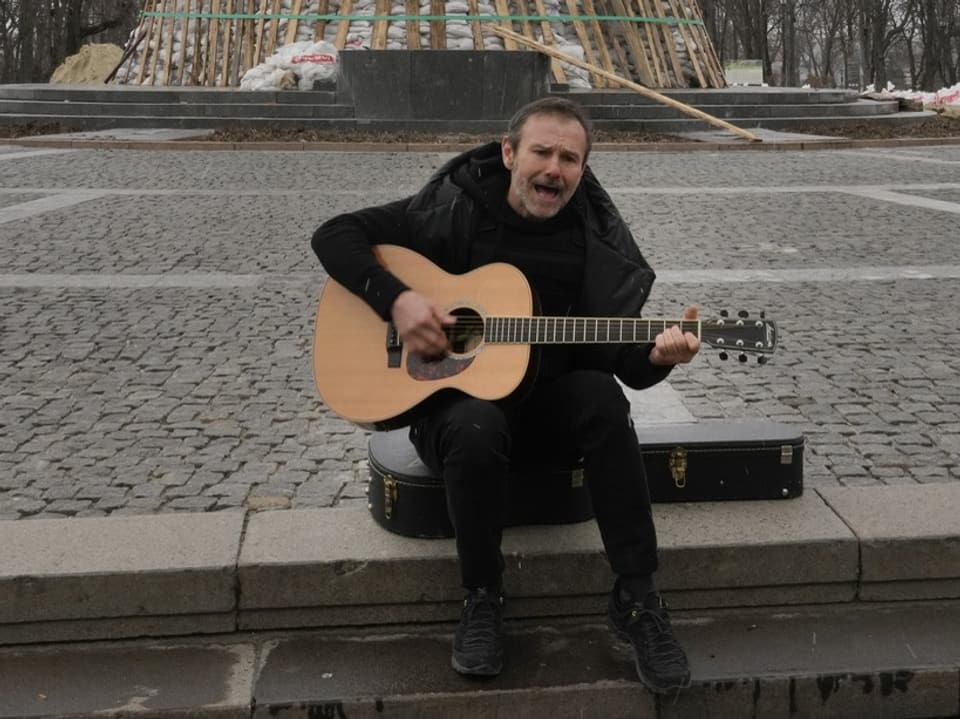 Mann spielt Akustikgitarre auf einer Parkbank.