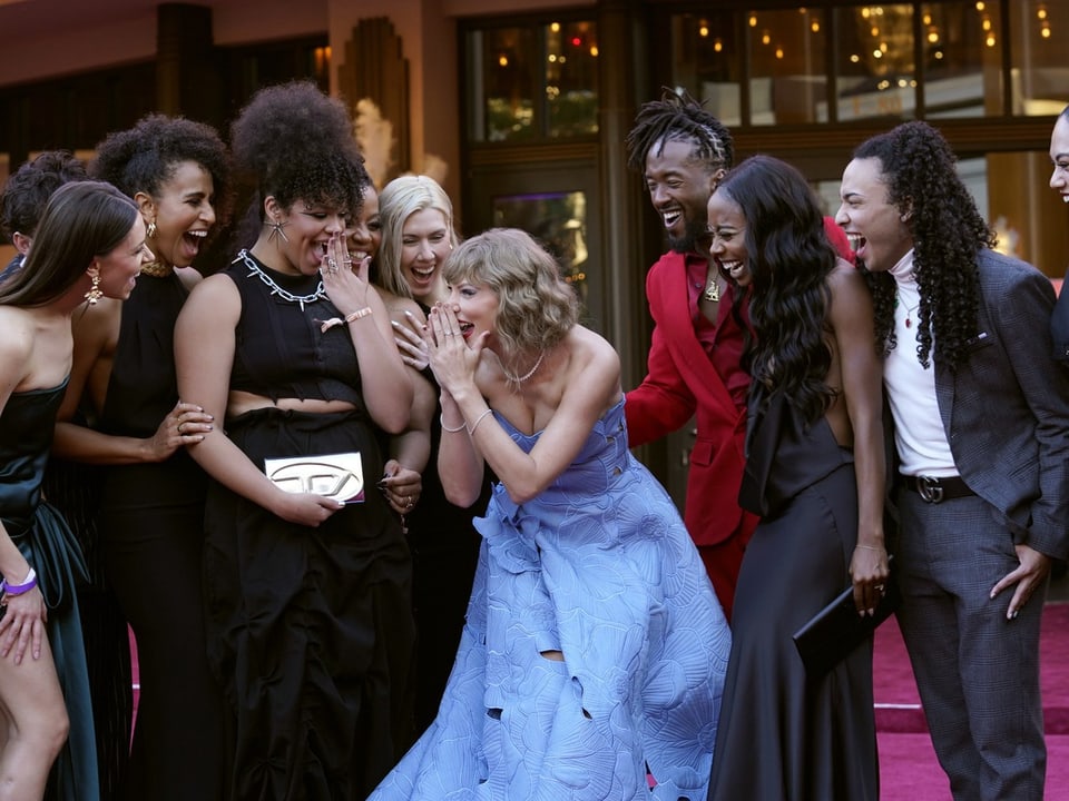 Taylor Swift in einem hellblauen Kleid auf dem roten Teppich zusammen mit ihren Bühnentänzerinnern und -tänzern.