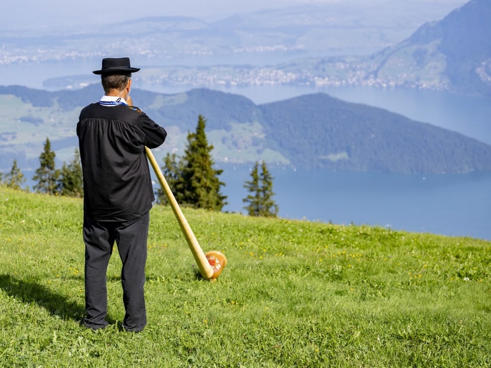 Mann spielt Alphorn mit Blick auf Berglandschaft und See.