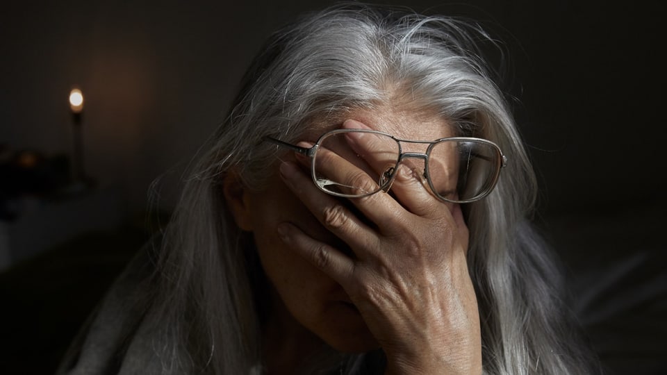 Eine ältere Frau mit langen grauen Haaren schiebt ihre Hand zwischen Brille und Augen und verdeckt damit ihr Gesicht.