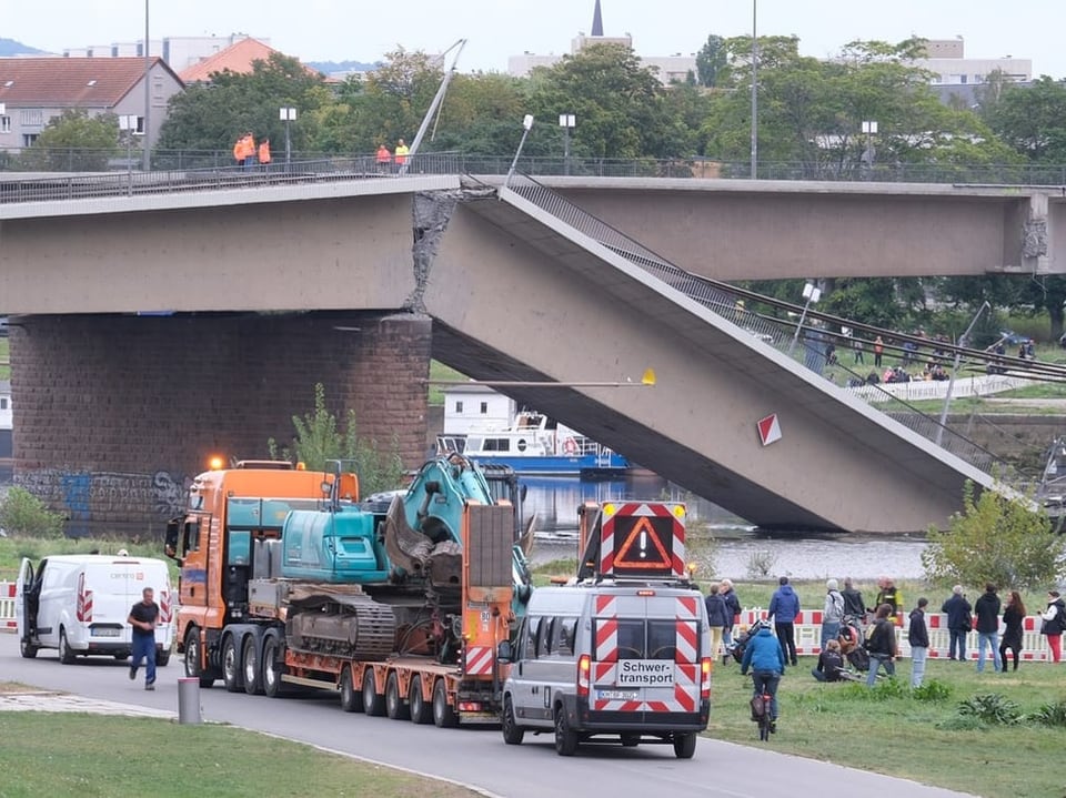 Eingestürzte Brücke über Fluss mit Schwerlasttransport und Baustellenfahrzeugen.