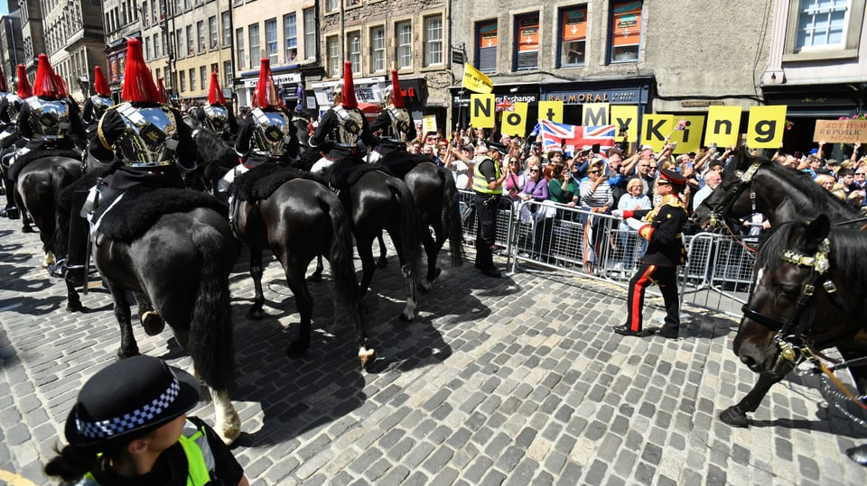 An der Parade laufen viele Pferde mit. Hinter der Zuschauerabsperrung halten Menschen Protestschilder hoch. 