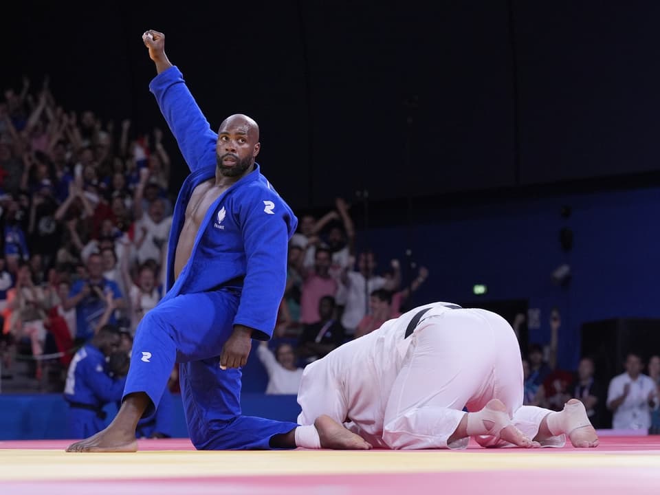 Judoka im blauen Gi feiert den Sieg, während Gegner am Boden kniet.