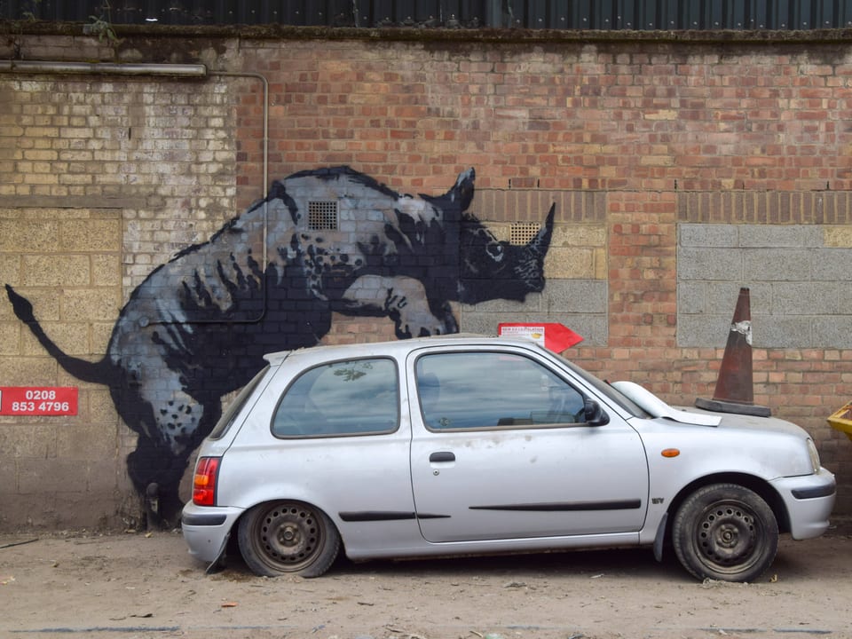 Graffiti eines Nashorns auf einer Ziegelmauer, das ein graues Auto scheinbar rammt.