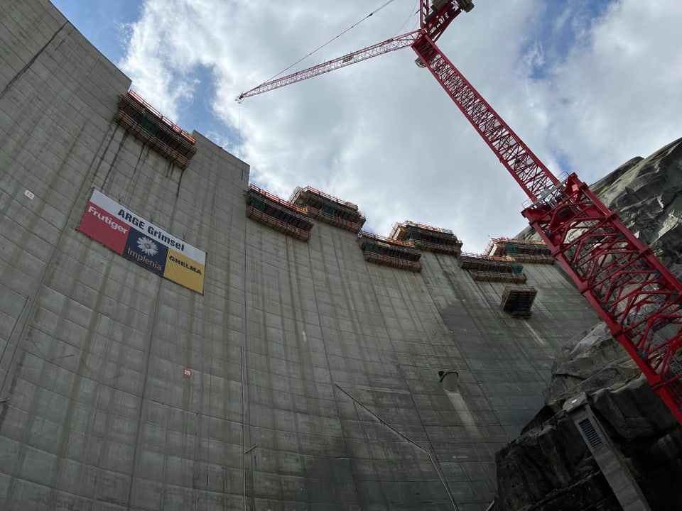 Betonmauer mit Baukran und Gerüsten vor bewölktem Himmel.