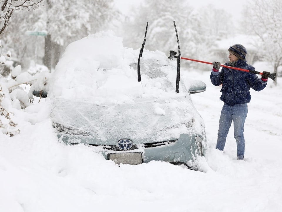 Eine Frau befreit ihr Auto mit Besen und Enteiser-Werkzeug vom Schnee. Es ist richtig gehend im Schnee eingesunken.