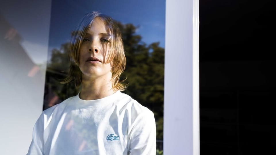 Person in weissem T-Shirt, mit halblangen blonden Haaren, schaut in Kamera.