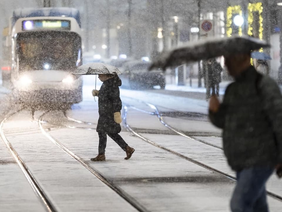 Menschen mit Regenschirmen überqueren schneebedeckte Strassenbahngleise.