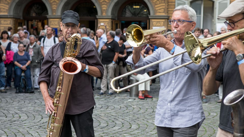 Drei Musiker mit Trompete, Posaune und Saxophon auf einem Dorfplatz. 