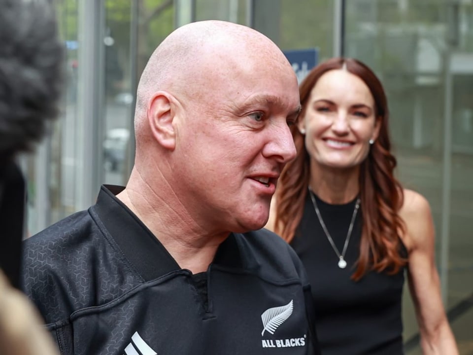 Zwei Menschen, einer im All Blacks-Trikot, lächelnd.