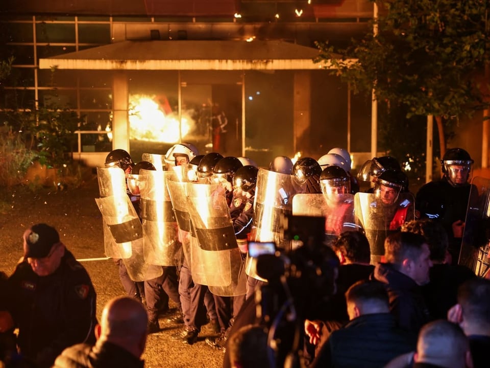 Polizisten mit Schilden vor einem brennenden Gebäude.