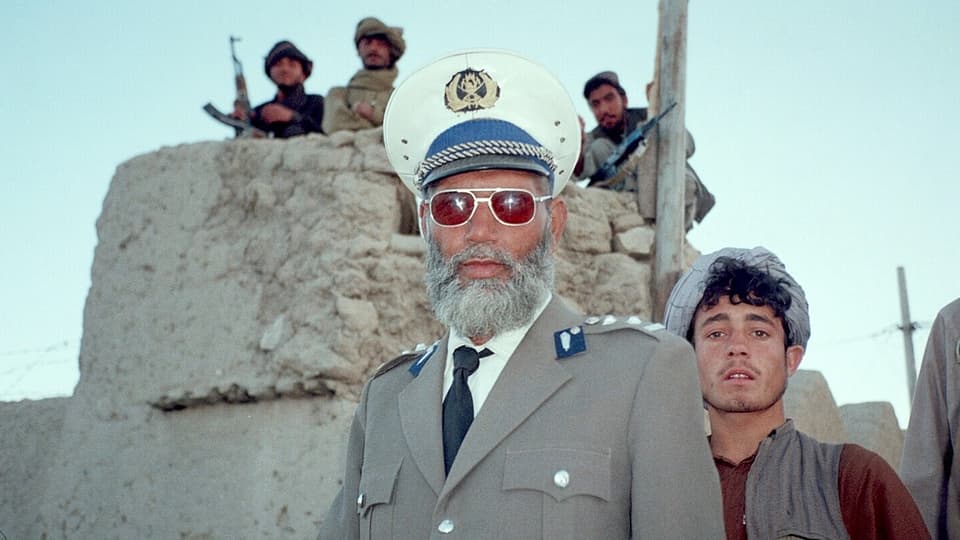 Ein Afghane mit Brille und Uniform blickt umgeben von bewaffneten Taliban-Kämpfern direkt in die Kamera.