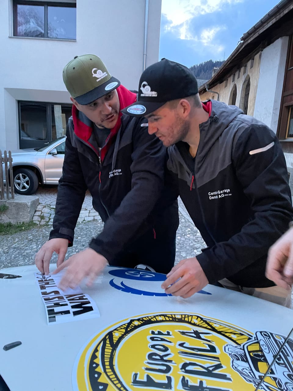 Peder Andri Janett und Orlando Bass kleben einen Kleber auf die Motorhaube.