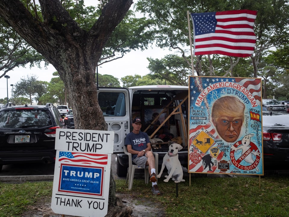 Mann mit Hund sitzt neben Trump-Postern und Flagge im Park.