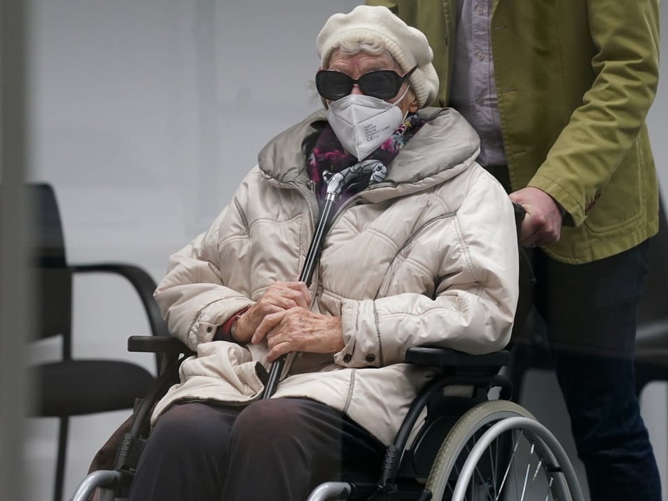 Ältere Person mit Maske in Rollstuhl sitzt drinnen.