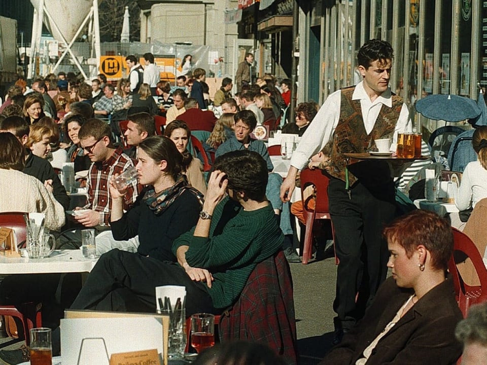 Voller Aussenbereich eines Cafés mit vielen Menschen und einem Kellner.