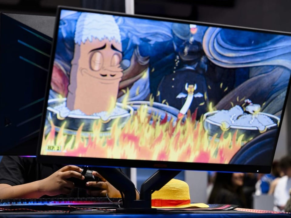 Ein Bildschirm, in dem ein Game zu sehen ist. Im Hintergrund sieht man jemanden, der das Game spielt.
