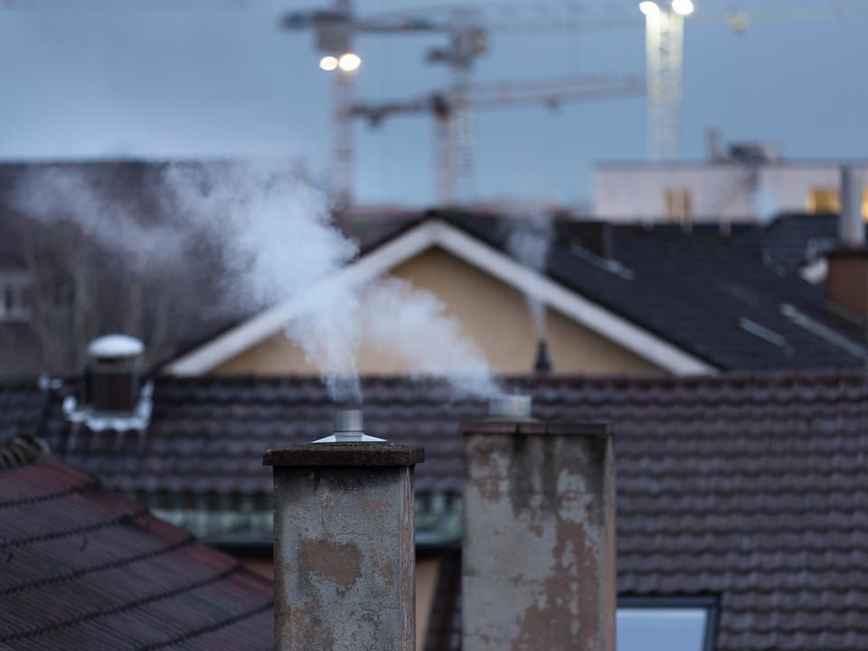Rauchender Schornstein vor einer städtischen Kulisse mit Kränen.