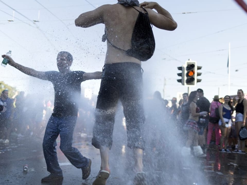 Zwei Personen tanzen vor einer Gruppe von Personen auf der Strasse und werden durch eine Wasserfontäne abgespritzt.