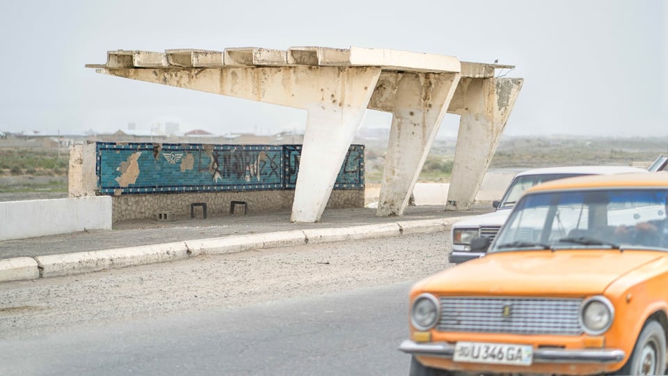 Eine Bushaltestelle aus weissen Betonpfeilern neben einer Strasse mit einem gelben Auto.