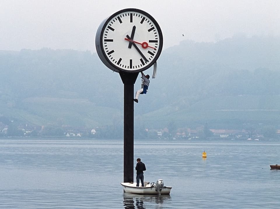 Zwei Personen an einer grossen Uhr im Wasser, eine kletternd, eine in einem Boot.