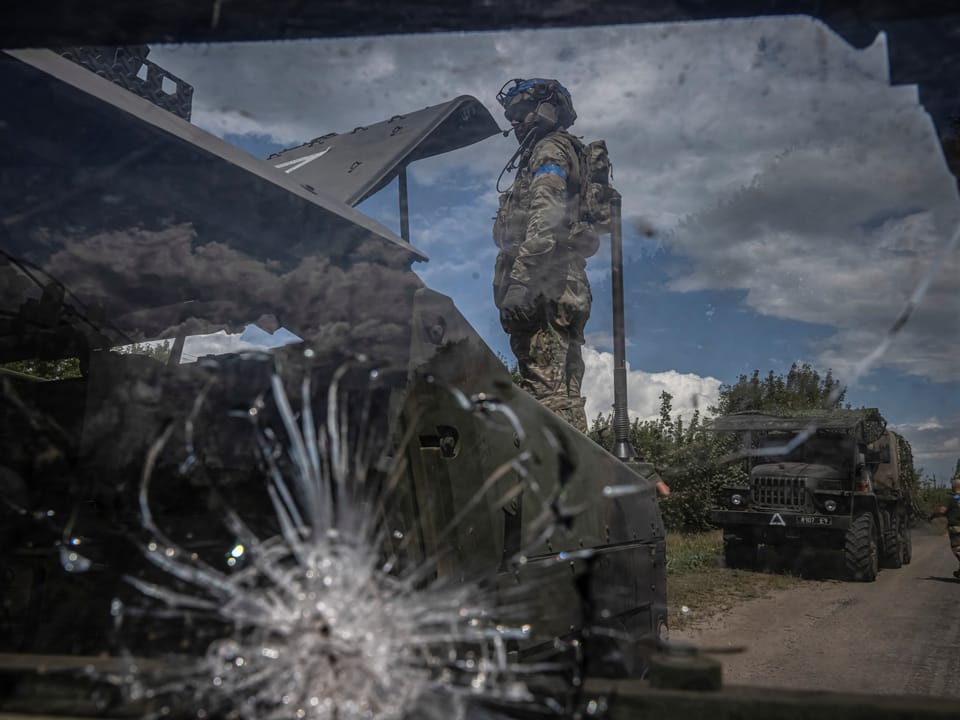 Soldat durch zerschossene Windschutzscheibe eines Fahrzeugs beobachtet, Militärfahrzeuge im Hintergrund.