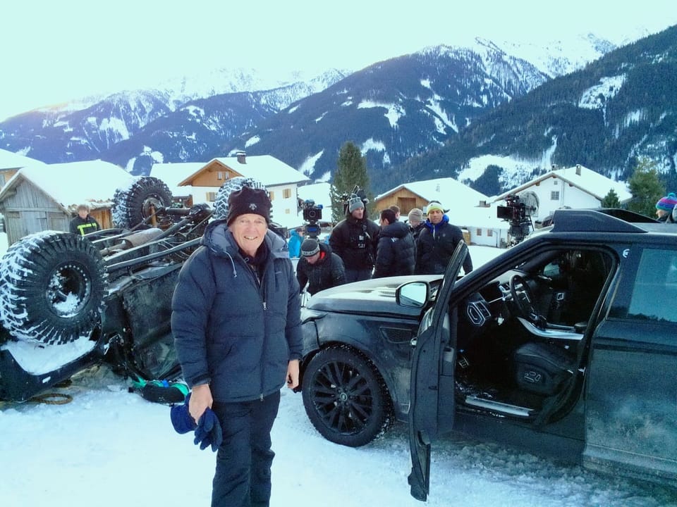 Mann vor einem schwarzen SUV und einem umgekippten Fahrzeug im Schnee, mit Bergen im Hintergrund.