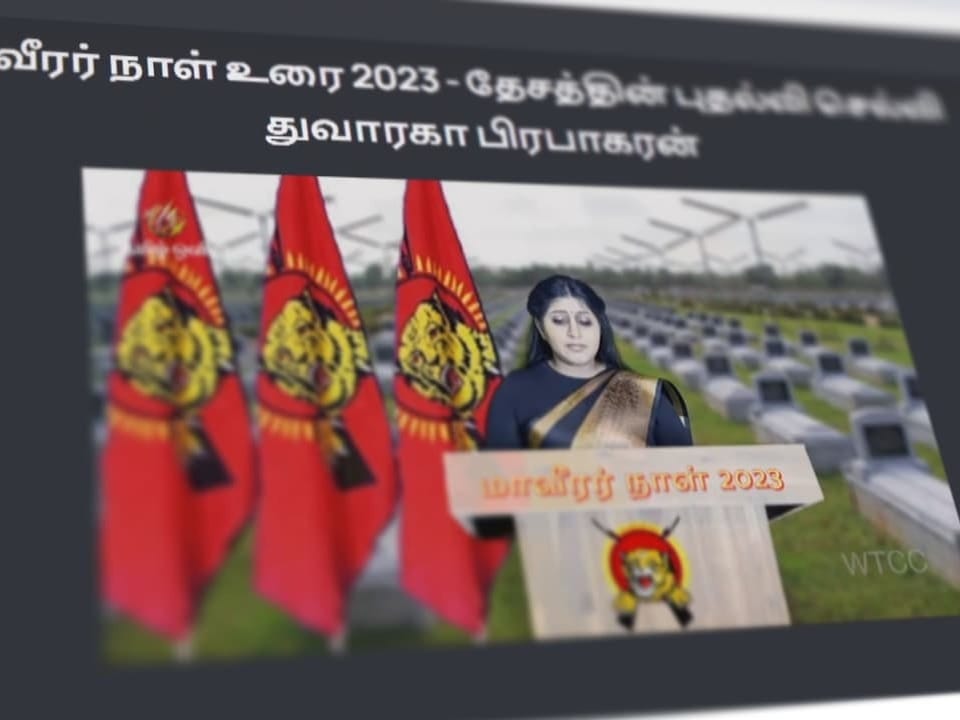 Frau hält Rede mit roten Flaggen und Gräbern im Hintergrund.