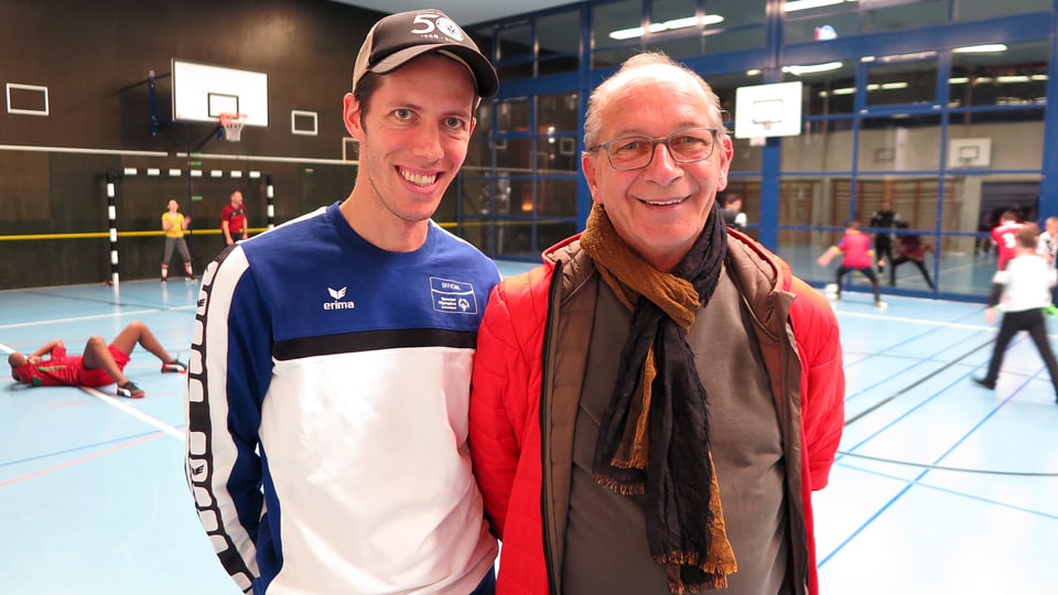 Zwei Männer in einer Turnhalle. Sie lächeln in die Kamera, im Hintergrund spielen Kinder Fussball.