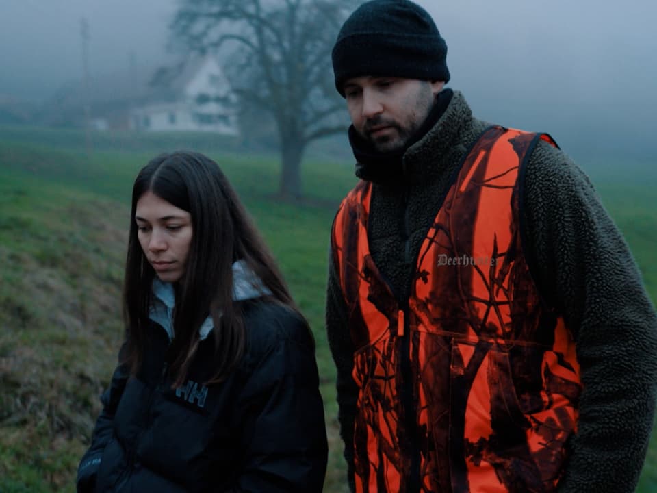 Alice und Dennis laufen nebeneinander im Wald. Alice trägt eine schwarze Jacke und Dennis eine orange Leuchtweste.