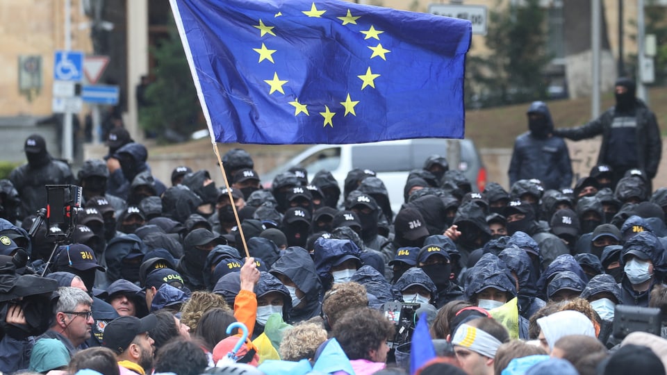 Protest von vielen Personen mit einer EU-Flagge. Vor ihnen stehen viele Polizeikräfte.