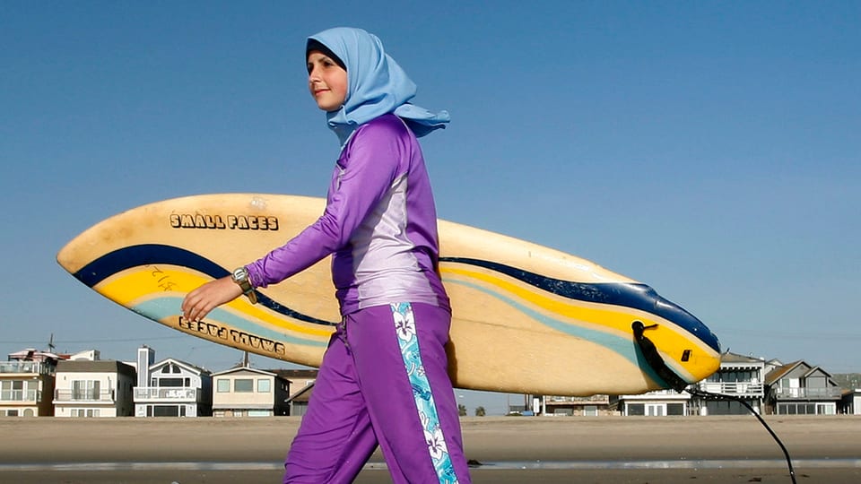 Eine Surferin in einem Burkini trägt ihr Brett an einem Strand.