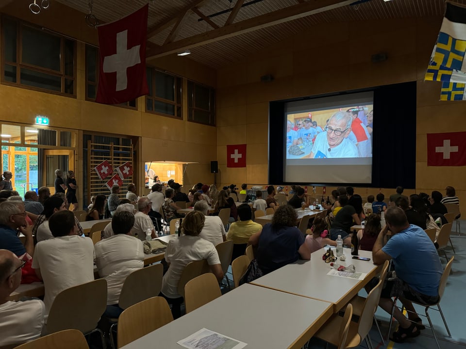 Menschen sitzen in einem Raum und schauen auf eine Projektionsleinwand.