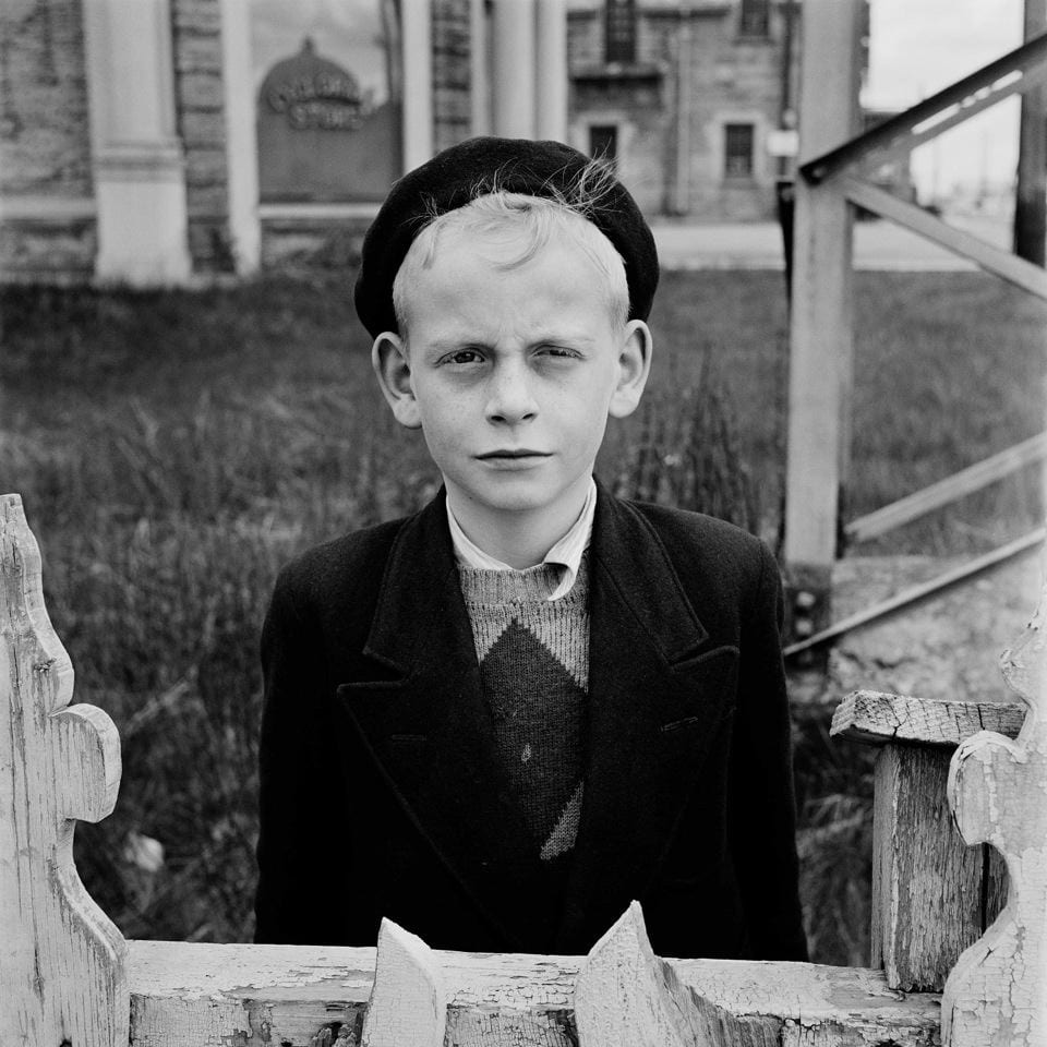 Ein Junge steht hinter einem Zaun. Er trägt einen Hut.