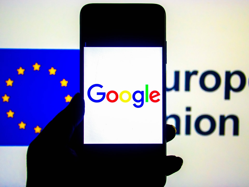 Eine schattierte Hand, die ein Smartphone hält, wo die Aufschrift Google draufsteht. Im Hintergrund ist die EU-Flagge.