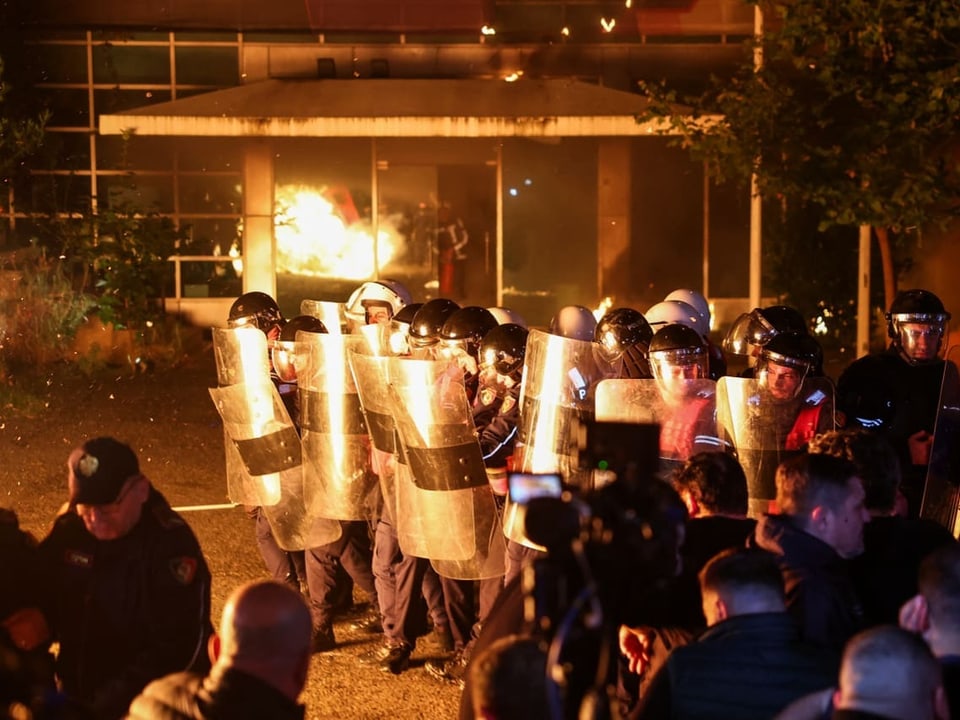 Polizisten mit Schutzschildern während eines Protests vor einem brennenden Gebäude.